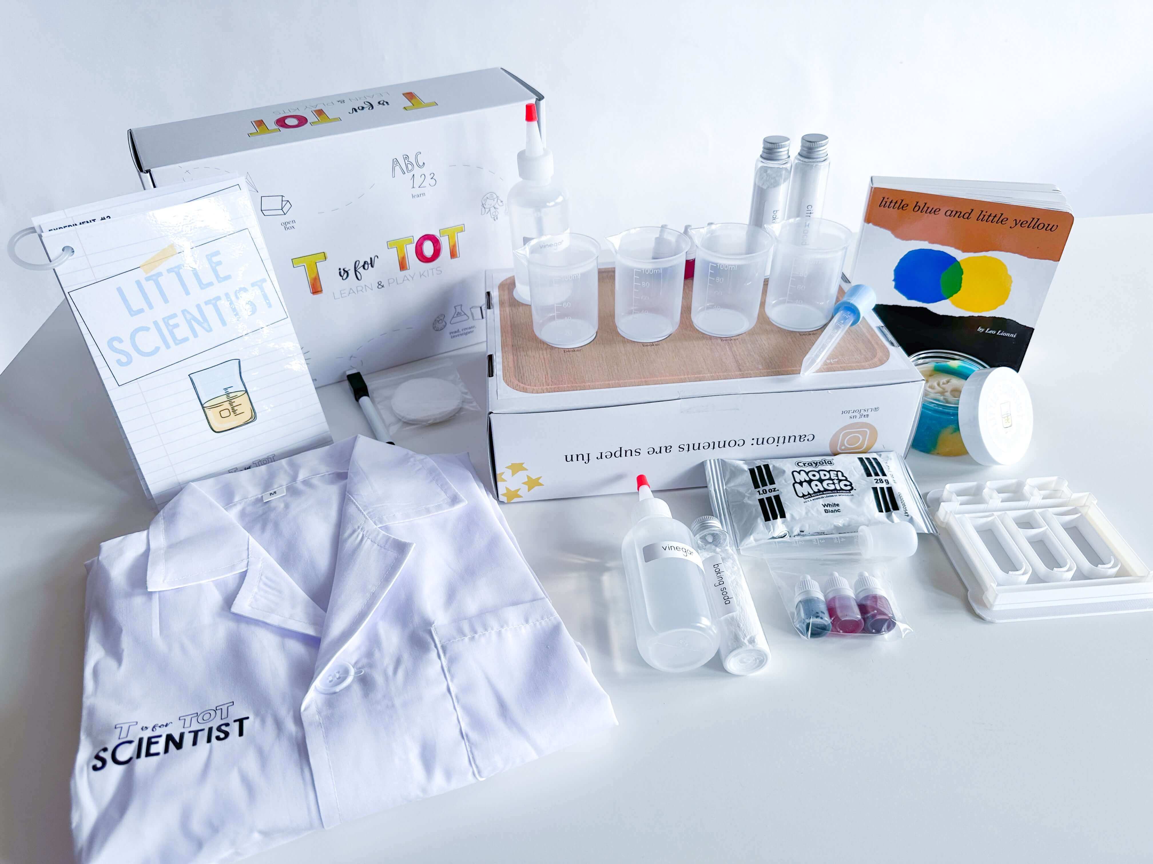 Little Scientist play kit for kids with beakers, pipettes, food coloring, and other science tools. Features a lab coat, experiment guide, and dry erase marker for interactive learning. Comes with homemade playdough, a lab playdough cutter, and 